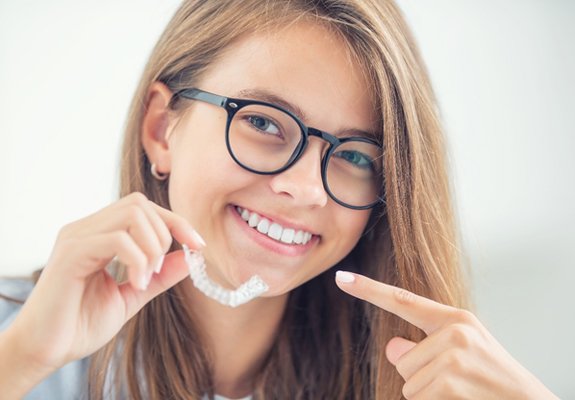 A young female wearing glasses and holding a clear aligner while pointing to her smile